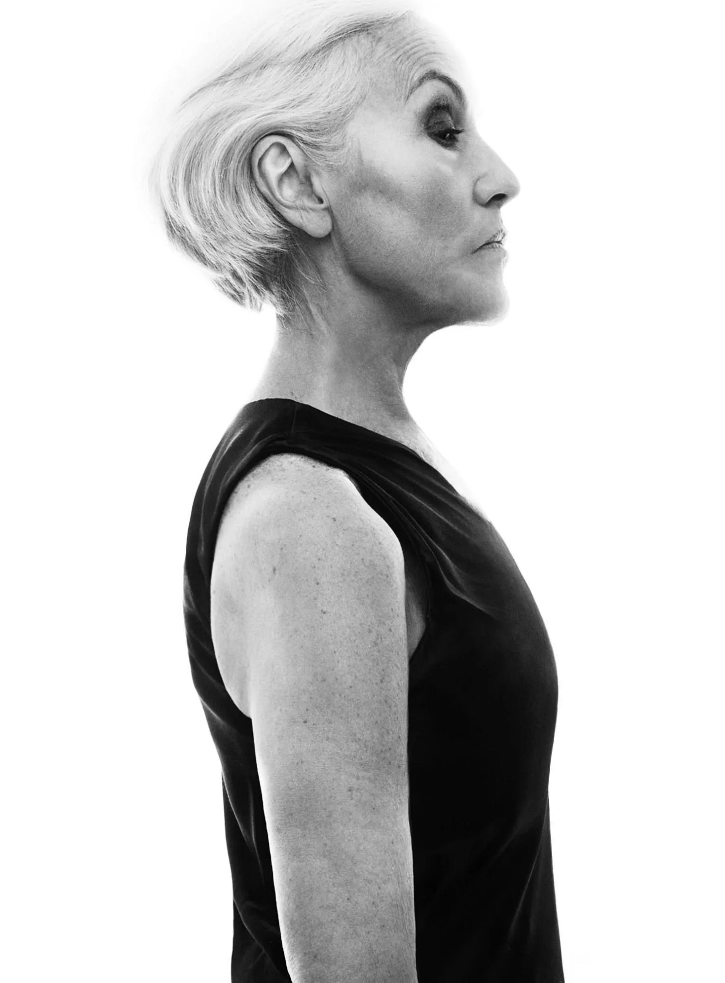 A woman photographed in black and white from the natural waist up stands in profile with a slicked back silver bob and a simple sleeveless black top.