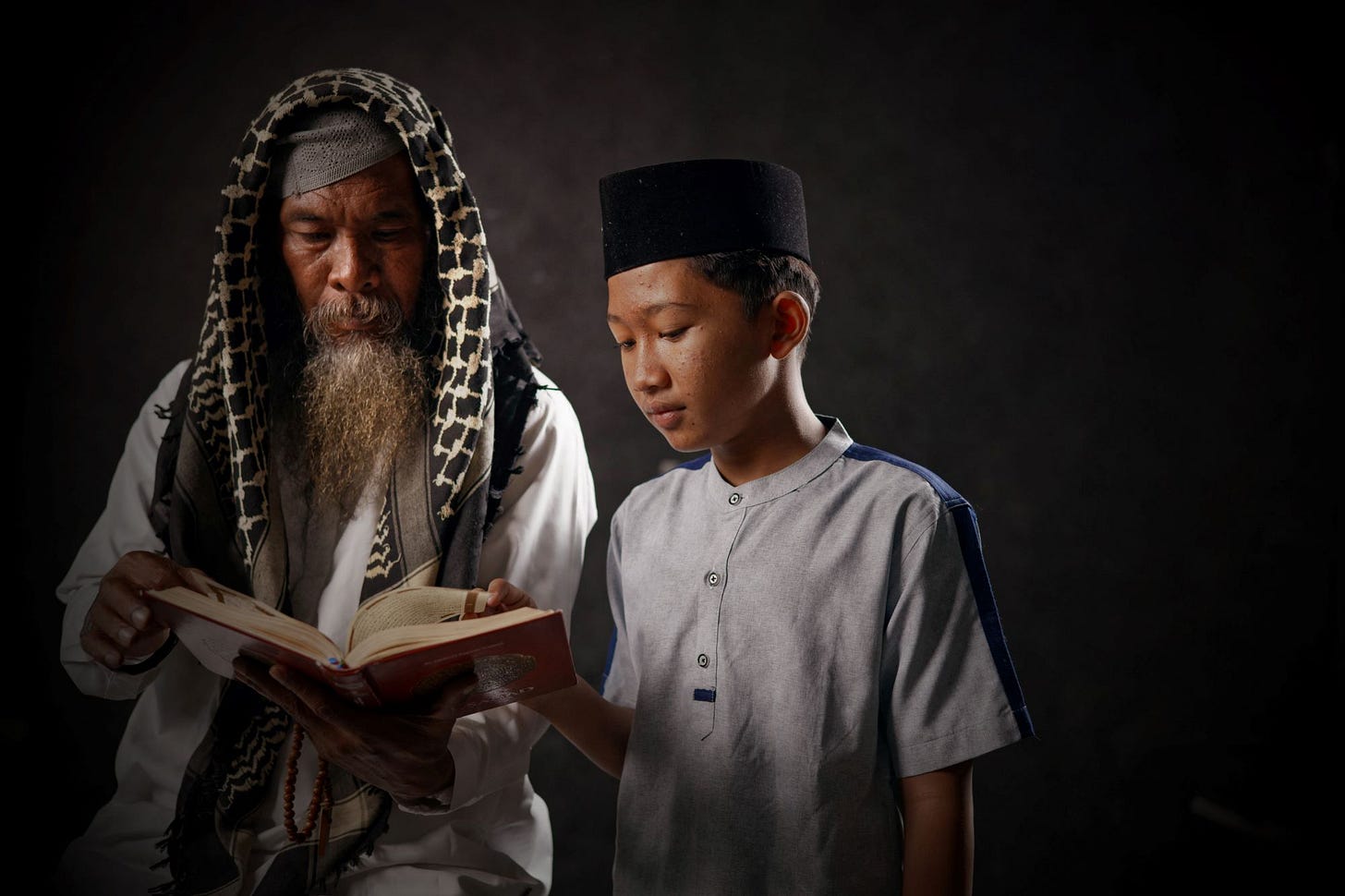 Bearded man in a keffiyah holding a book and instructing a youth 