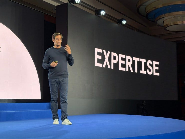 A speaker on stage presenting at the Checkout.com conference, with the word 'EXPERTISE' displayed prominently in the background.