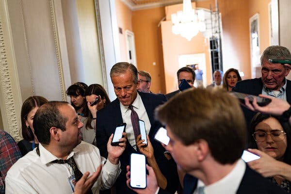 Senator John Thune, wearing a dark suit, is surrounded by people holding phones in his direction. Senator John Thune, wearing a dark suit, is surrounded by people holding phones in his direction.