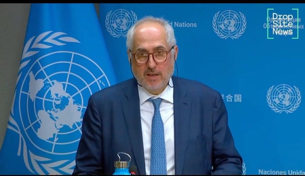 A man with glasses and balding hair wearing a dark suit white shirt and blue tie stands at a podium holding a blue water bottle. Behind him are multiple blue United Nations flags and a blue backdrop with the United Nations logo. A Drop Site News watermark appears in the upper right corner.