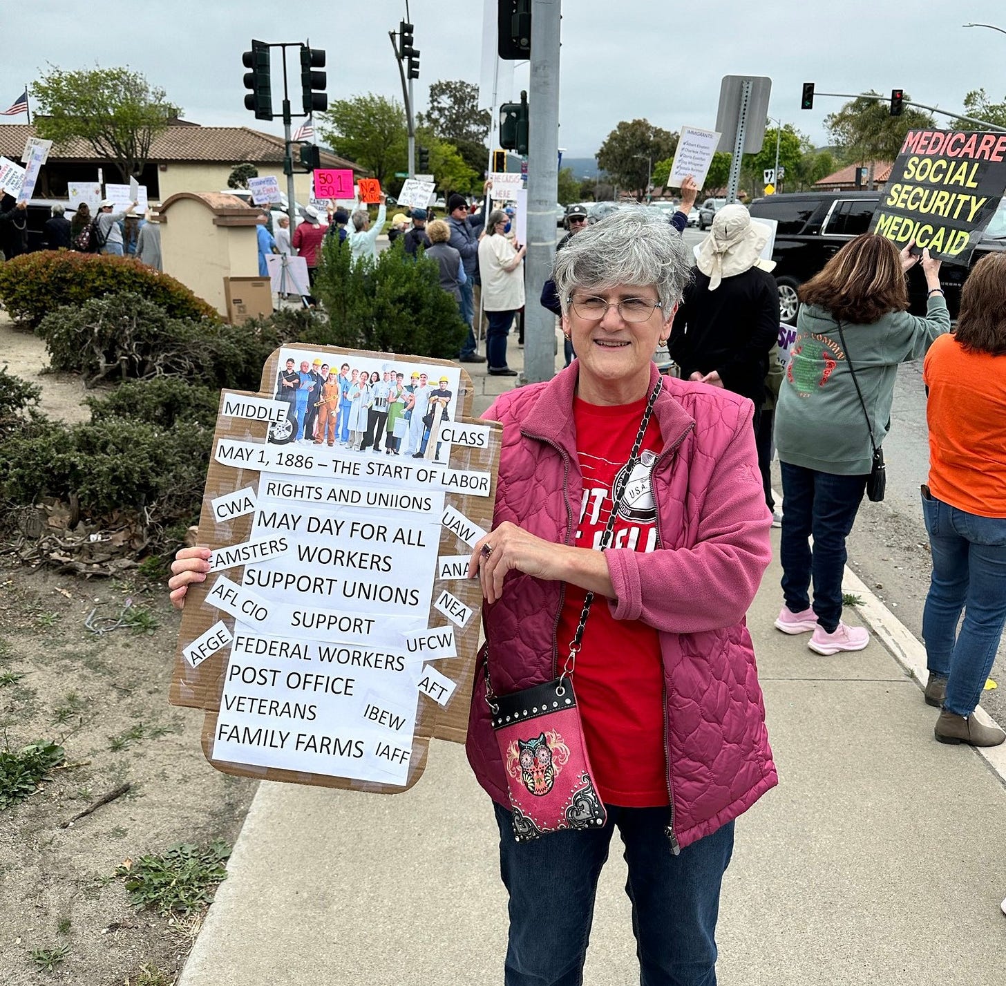 Protesters crowd around a street corner, mostly with their backs to the camera. They hold signs includeding one that reads "50501," another that reads "No Kings," another that reads "The Crisis is Here," and one that reads "Medicare, Social Security, Medicaid." In the foreground, a woman in a Fight Like Hell shirt faces the camera with a cardboard sign. It has pictures of working people in various work clothes, and reads "Middle Class - May 1, 1886, the Start of Labor Rights and Unions. May Day for All Workers. Support Unions, SUpport Federal Workers, Post Office, Veterans, Family Farms, IBEW, IAFF, AFT, UFCW, NEA, UAW, ANA, CWA, TEamsters, ALF-CIO, AFGE."