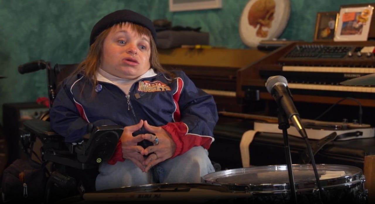 A disabled woman is giving an interview behind a drum set.