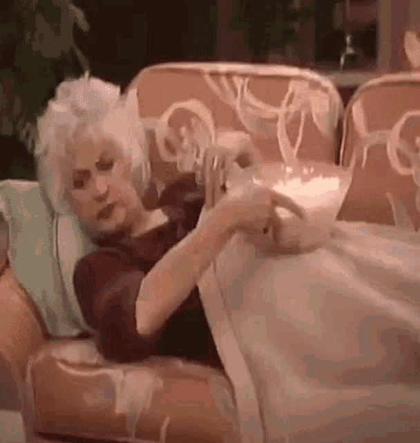 an elderly woman is laying on a couch with a bowl of popcorn .