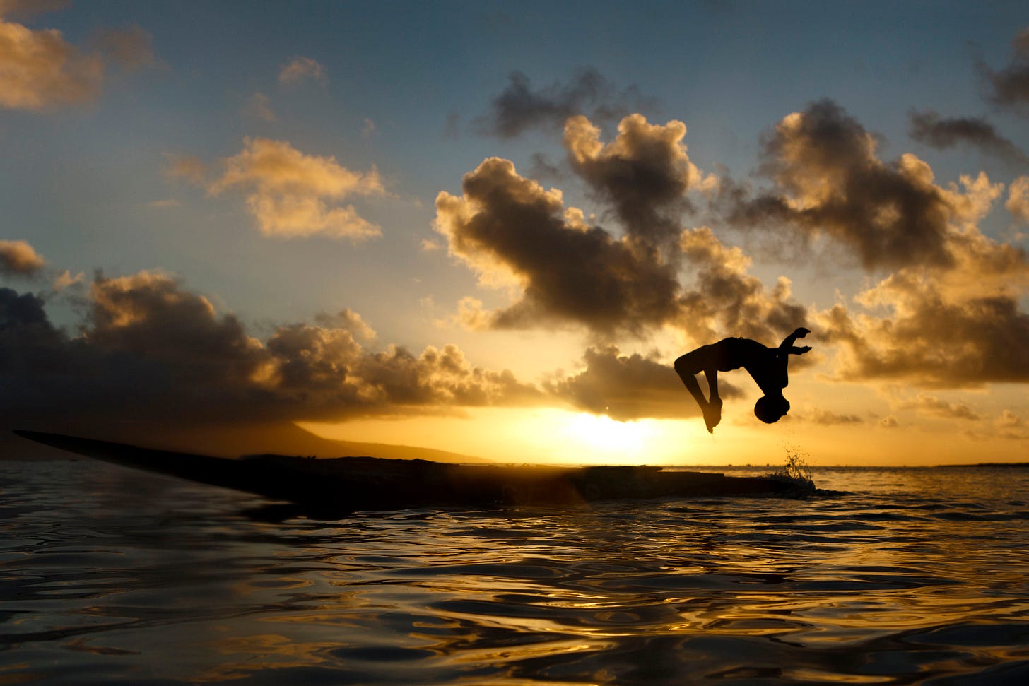 A person performs a backflip off a boat into the water at sunset, silhouetted against a dramatic sky with clouds and golden sunlight reflecting on the surface.