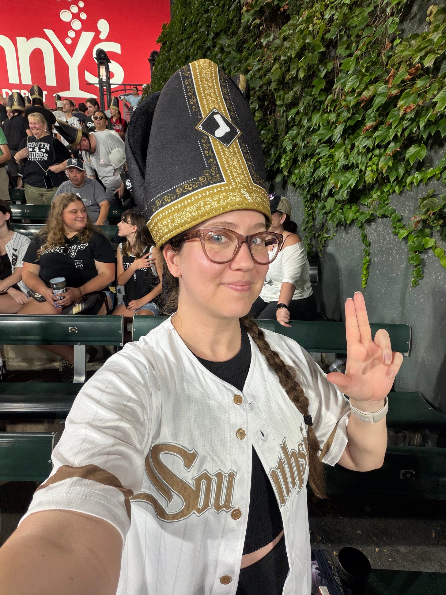 Woman in a White Sox jersey and glasses wearing a novelty Pope hat with a White Sox logo on it; behind are bleachers full of other baseball fans and an ivy-covered wall Woman in a White Sox jersey and glasses wearing a novelty Pope hat with a White Sox logo on it; behind are bleachers full of other baseball fans and an ivy-covered wall