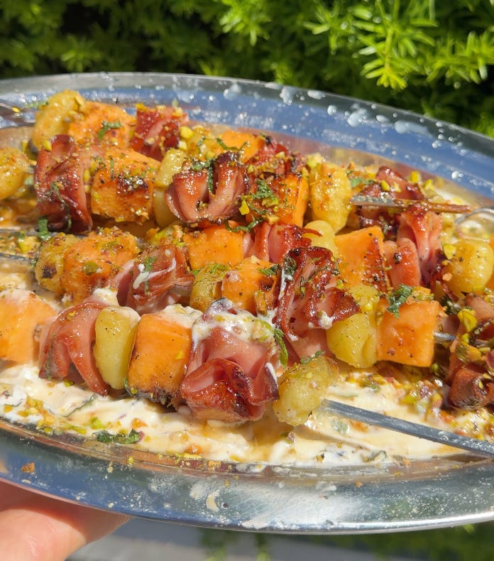 hand pulling bbqd mortadella skewers with crispy gnocchi on a silver platter over whipped ricotta and pistachio crumble with hot honey