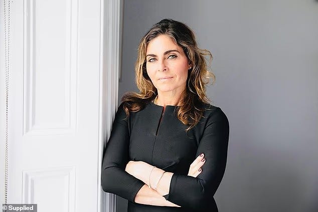Emma-Jane Taylor stands indoors with her arms crossed, leaning against a white doorframe. She wears a black long-sleeve dress and looks at the camera against a pale gray wall.