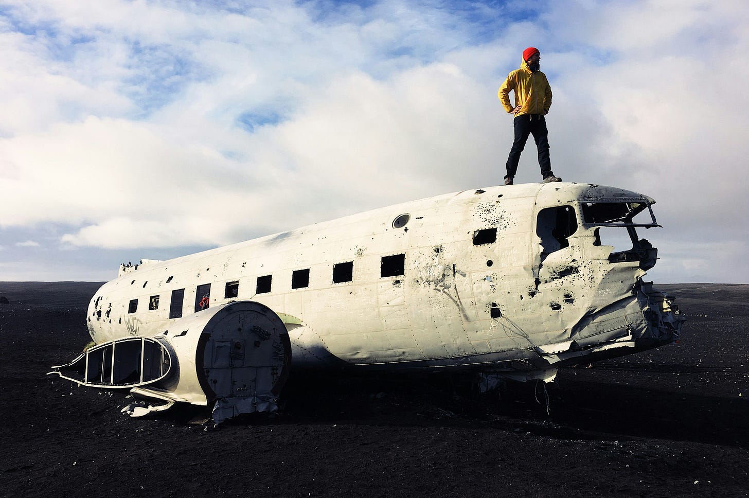 Person in yellow jacket and red beanie standing on top of a crashed white airplane wreckage on black volcanic sand under a cloudy sky Person in yellow jacket and red beanie standing on top of a crashed white airplane wreckage on black volcanic sand under a cloudy sky