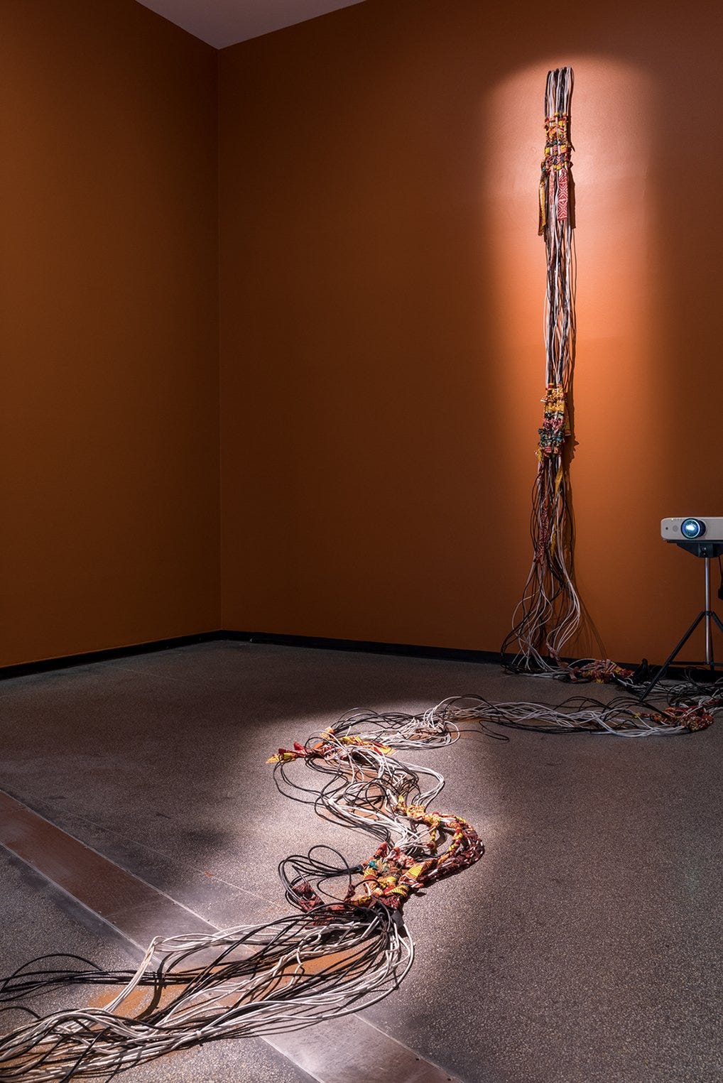 A bunch of cables pinned to a wall and flowing down towards the floor in a gallery with an ochre wall in the background and spotlight trained on the top half of the cables. At intervals, the cables are tied together with colourful African wax print cloth. A bunch of cables pinned to a wall and flowing down towards the floor in a gallery with an ochre wall in the background and spotlight trained on the top half of the cables. At intervals, the cables are tied together with colourful African wax print cloth.
