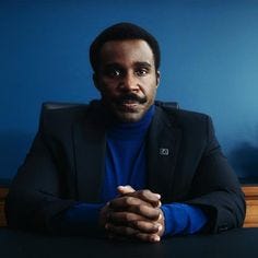 This may contain: a man sitting at a table with his hands folded and looking off to the side