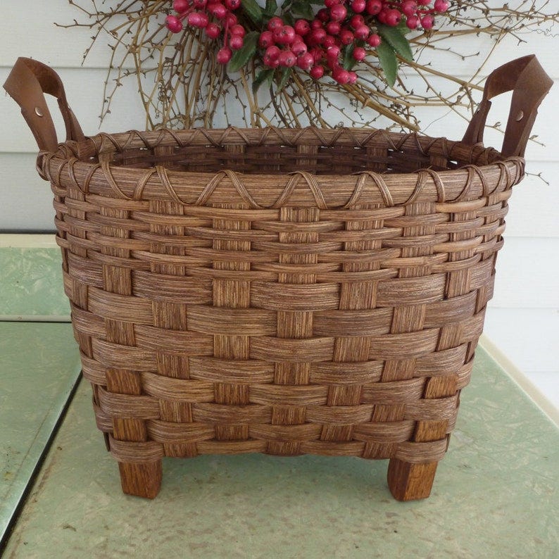 May include: A brown woven basket with two leather handles and four wooden legs. The basket is oval shaped and has a woven pattern.