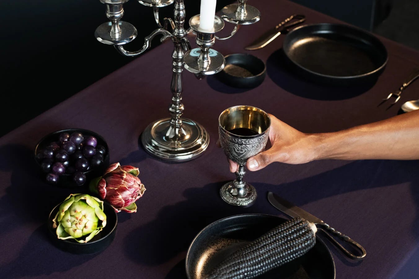A imagem mostra uma mesa de jantar com cores escuras e sombrias, um candelabro ornamentado e uma taça de vinho prateada.