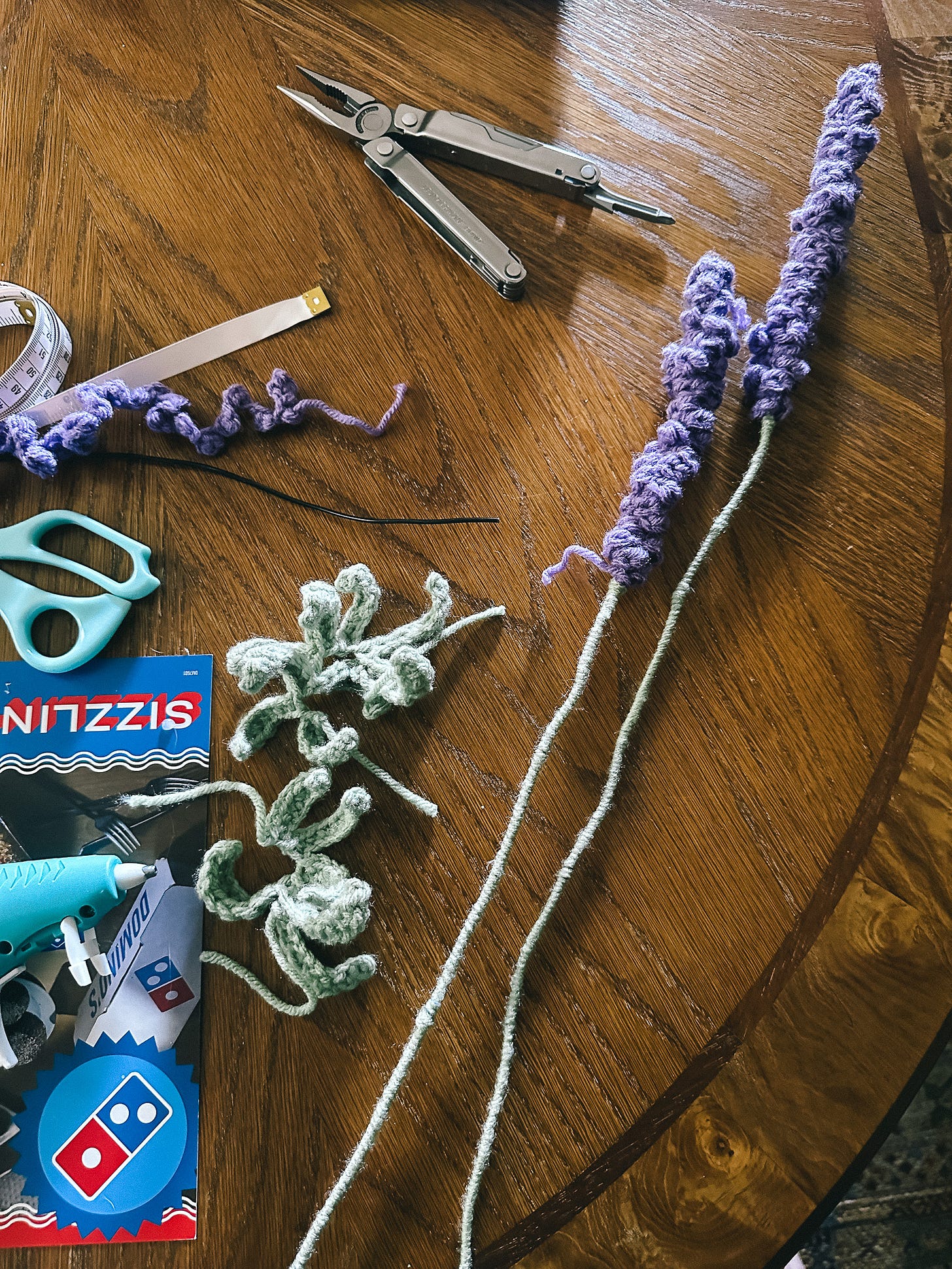 aerial view of craft supplies on a wooden table, with two lavender flowers made of yarn in the process of being hot glued