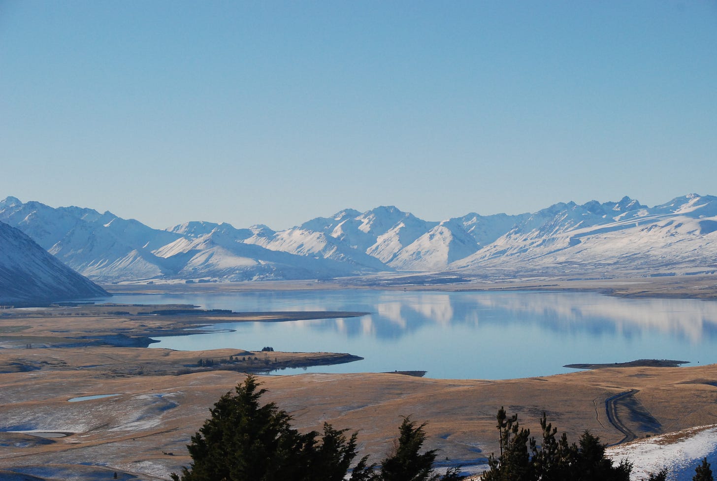 Photograph of Lake Tekapō in winter from the Mt John Observatory. The sky is light blue and clear, the mountains in the distance are covered with snow, tops of trees are visible in the foreground.