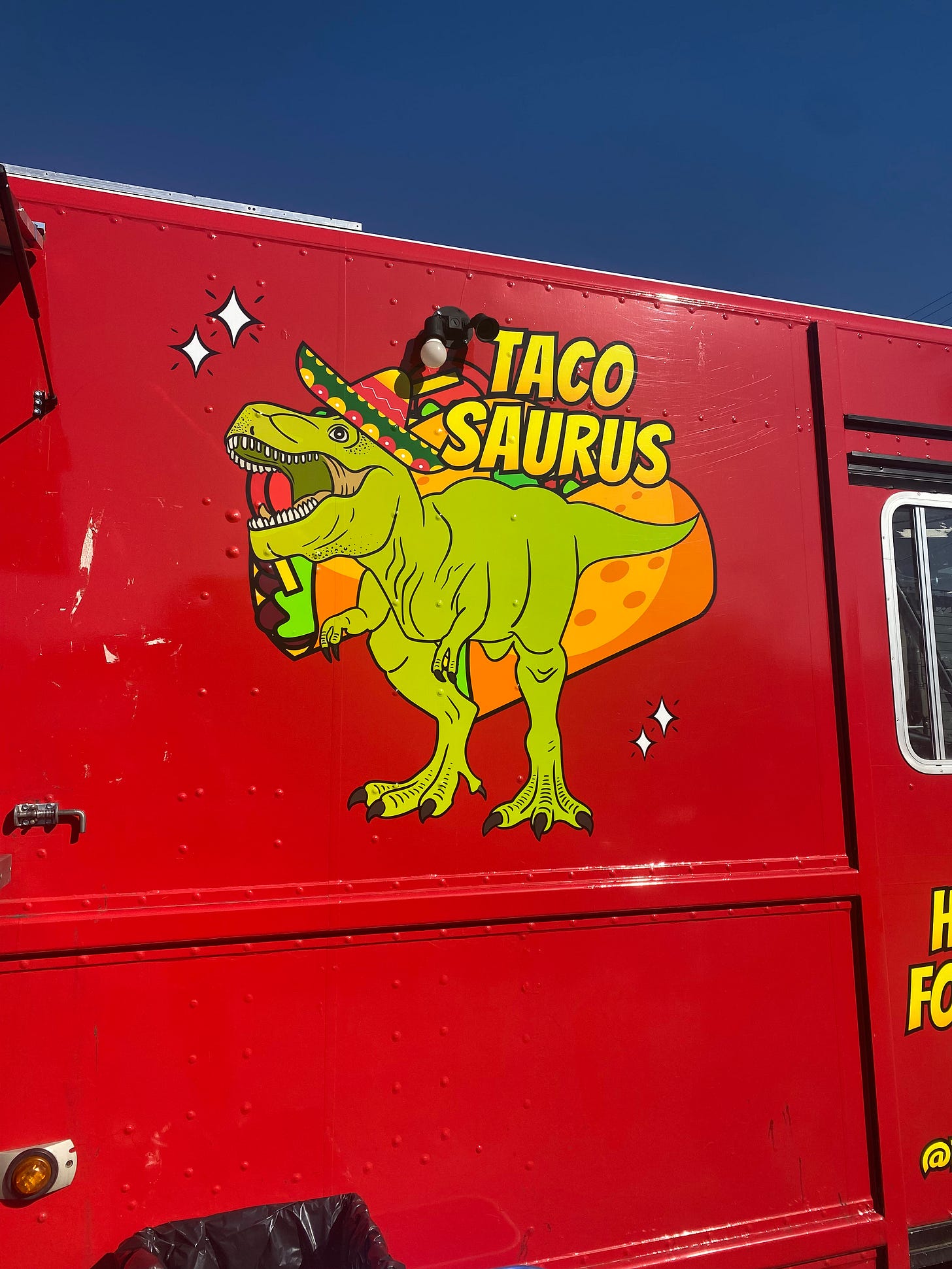 A red food truck side panel that reads 'TACOSAURUS' and obviously, has an illustration of a t-rex in a sombrero.