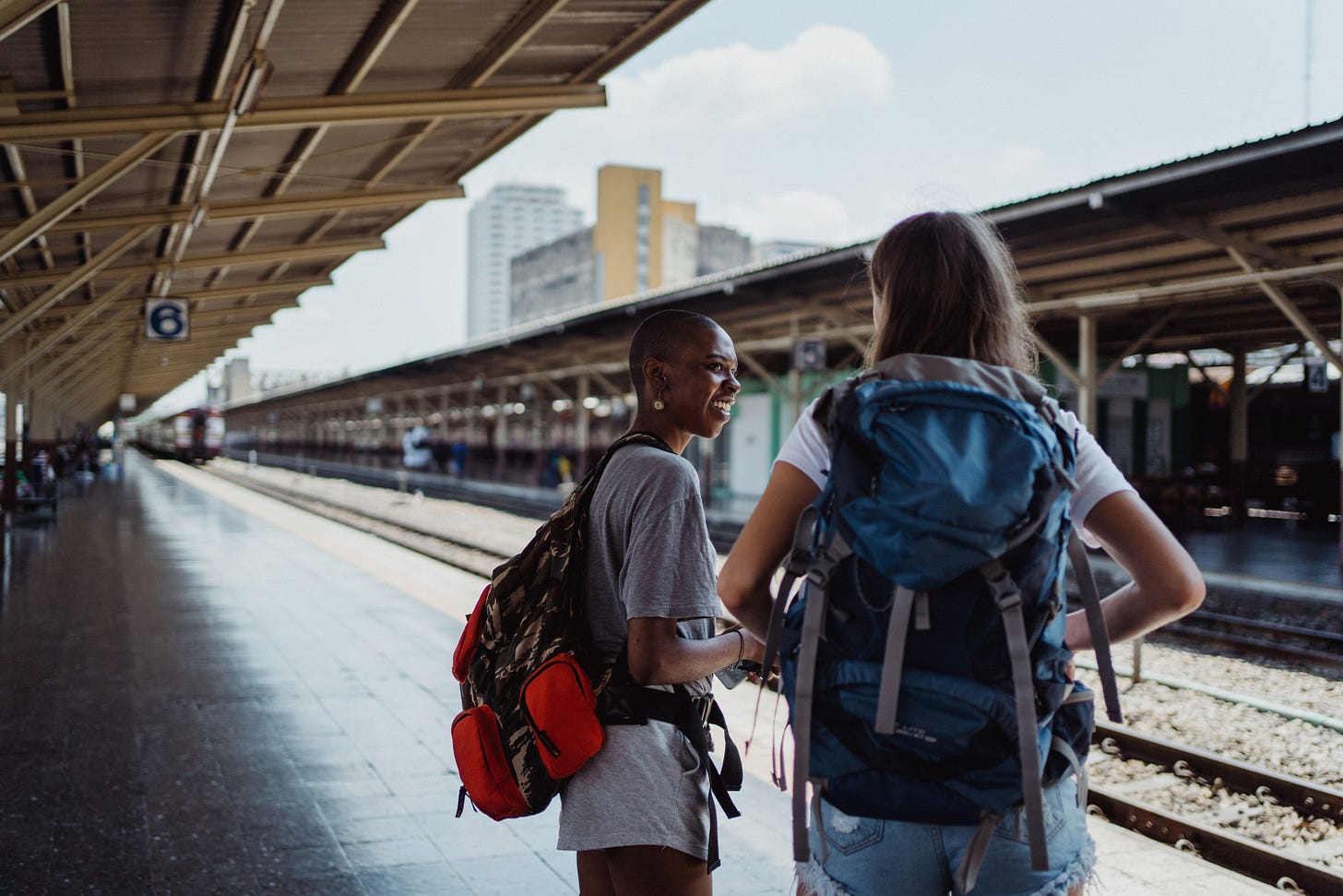 Two female backpackers wait on a sunny railway platform, ready for adventure.