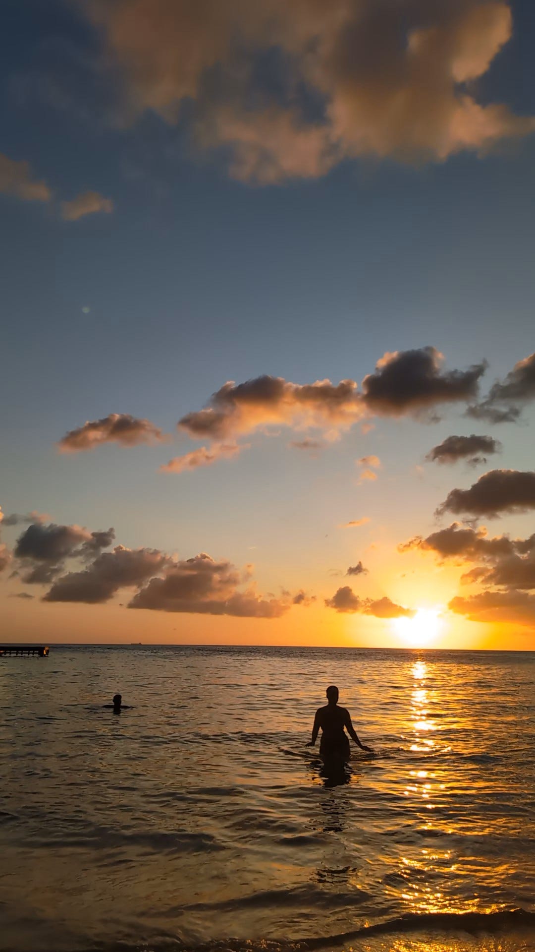 Sunset over the ocean with golden light reflecting across the water. Two people stand in shallow water as silhouettes, facing the horizon. Soft clouds glow orange and pink against a fading blue sky, and a small pier extends from the left side of the frame. Sunset over the ocean with golden light reflecting across the water. Two people stand in shallow water as silhouettes, facing the horizon. Soft clouds glow orange and pink against a fading blue sky, and a small pier extends from the left side of the frame.