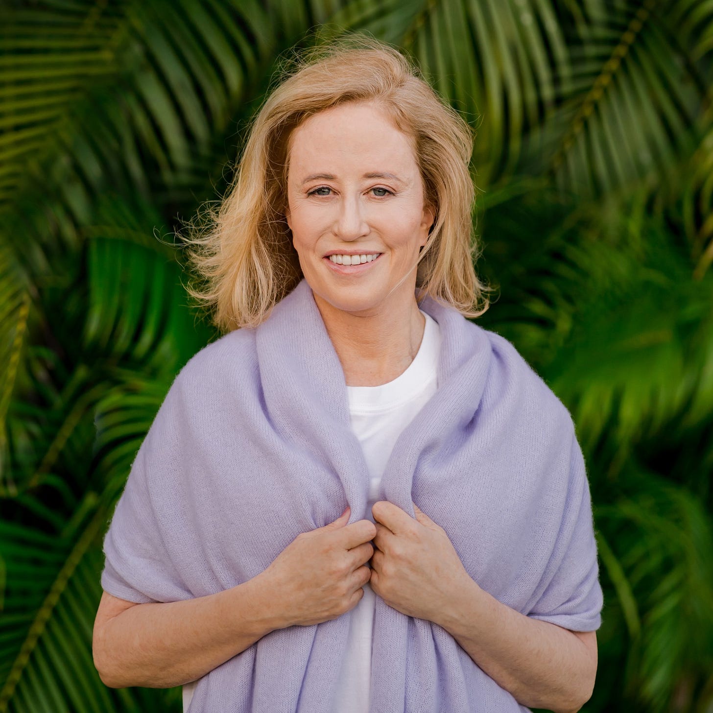 Image of Kim Samuel, wearing a purple pull over and a white shirt, smiling broadly at the camera, with green plants in the background.