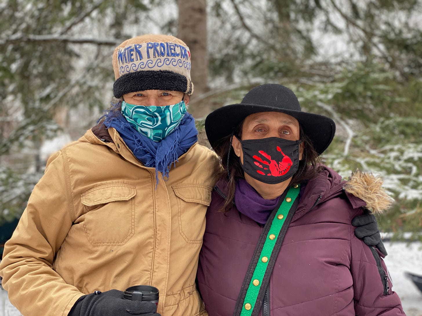 Marian and Winona looking at the camera, Marian's arm around Winona, both are wearing big puff coats, hats and bandanas over their mouths and most of their faces. It is winter and there are trees and snow in the background. Winona's face covering has a red hand print on the front. Marian's hat says 