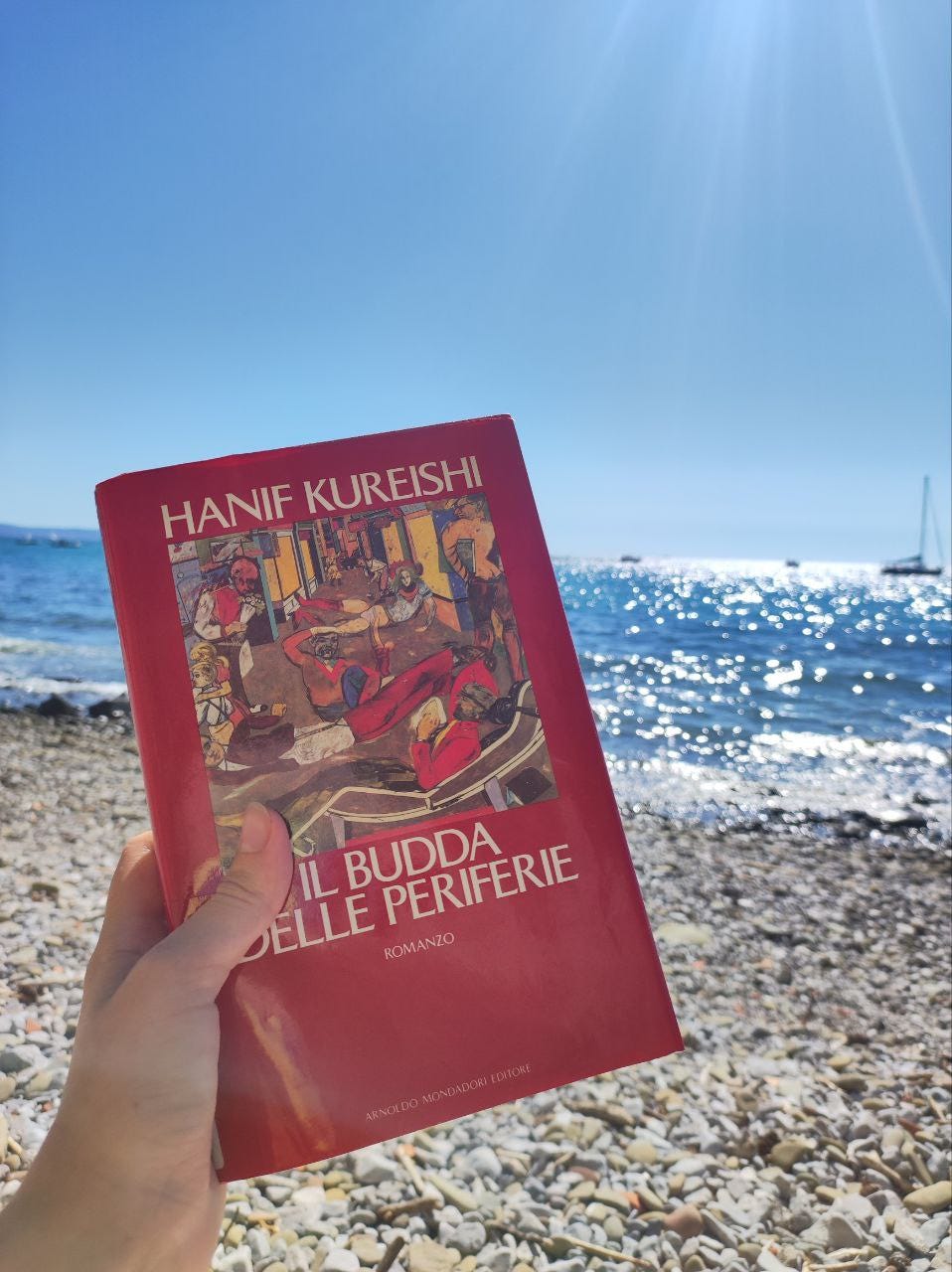 Foto al mare. Sullo sfondo si vede una spiaggia e il mare, azzurro. è una giornata di sole splendente e il cielo è senza una nuvola. C'è anche una barca a vela all'orizzonte. In primo piano, una mano tiene un libro. Si tratta di Il budda delle periferie, di Hanif Kureishi. La copertina è bordeaux con un quadro grande che occupa parte della copertina. 