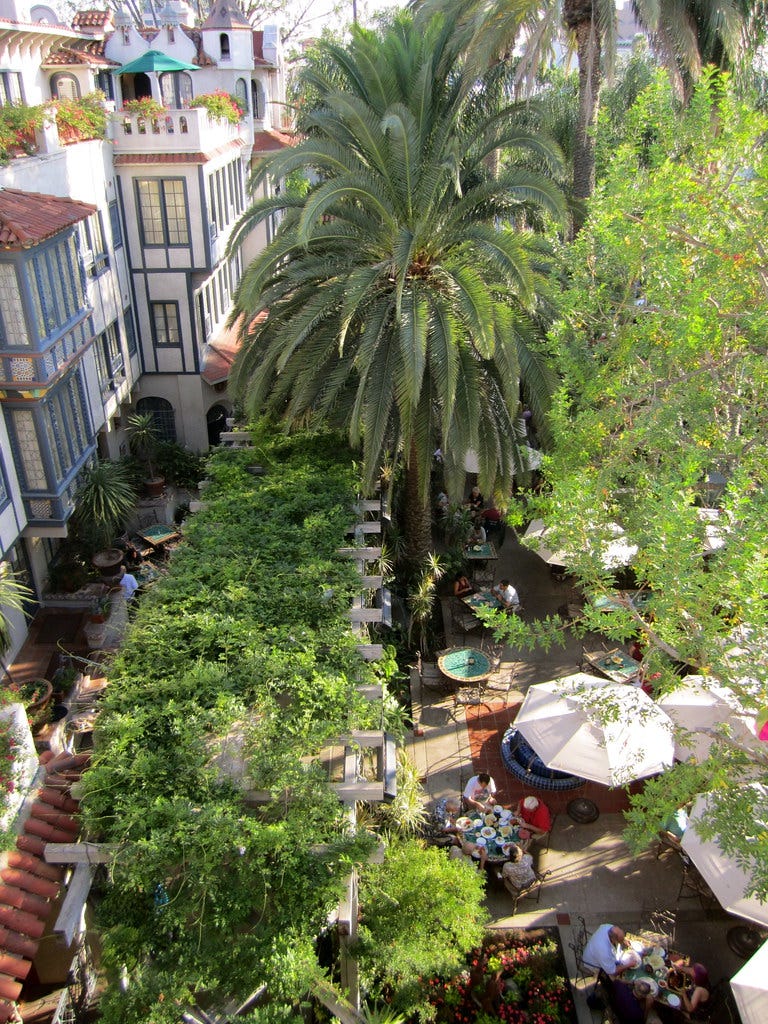 photo of a courtyard cafe shaded by trellised vines and a palm tree. People are sitting around small colorful tables eating and drinking. The buildings around the courtyard have friendly-looking windows and balconies. photo of a courtyard cafe shaded by trellised vines and a palm tree. People are sitting around small colorful tables eating and drinking. The buildings around the courtyard have friendly-looking windows and balconies.