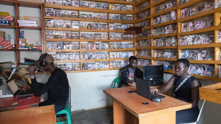 A photo of three people on computers inside an internet cafes with dvds for sale on the walls behind them.