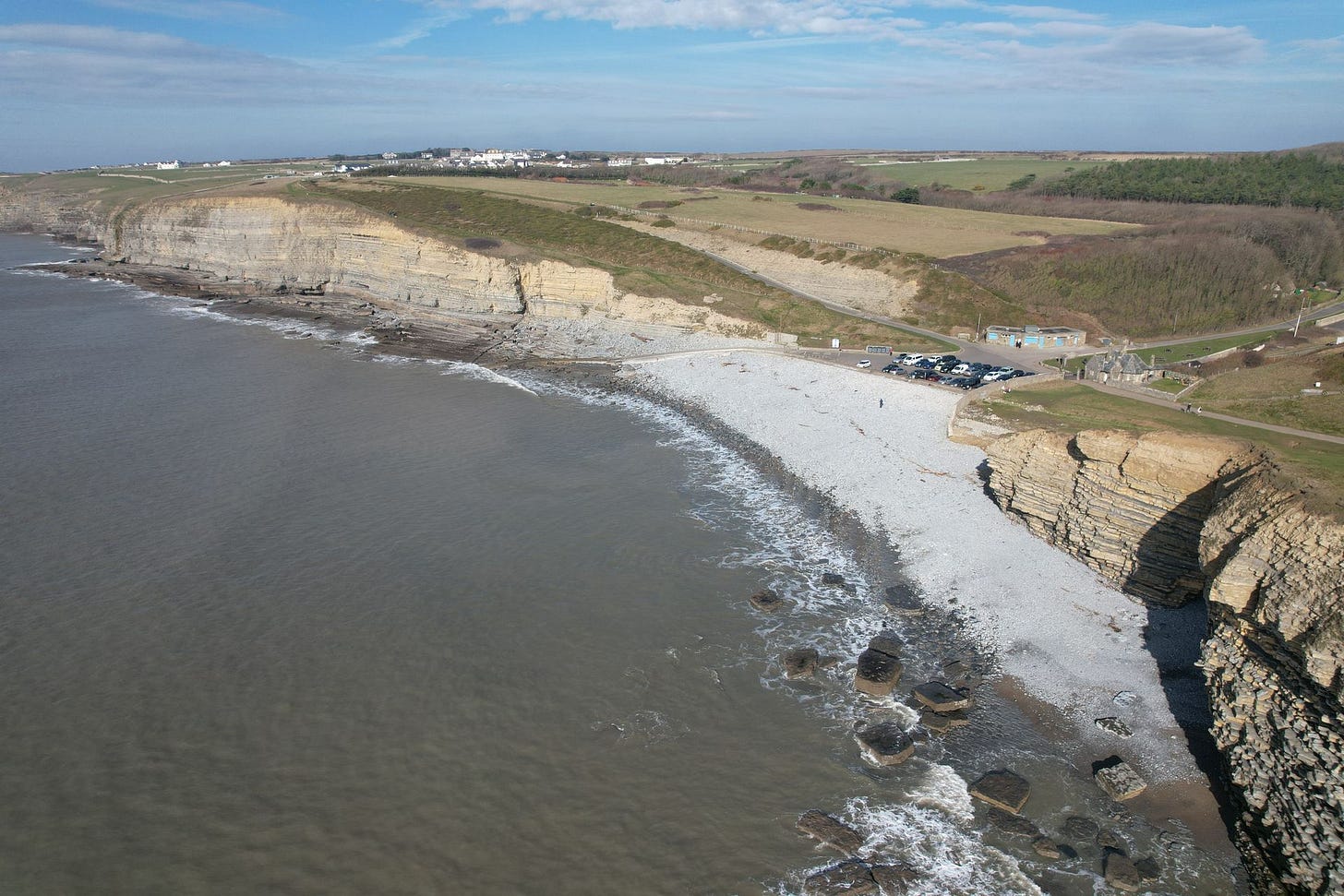 An aerial view of a curving coastline featuring steep, horizontally layered rock cliffs dropping down to a light grey pebble beach. The ocean water washes over submerged rocky platforms in the foreground. Above the cliffs lies flat green farmland, and a car park with several vehicles and a small building is visible nestled just above the shoreline.