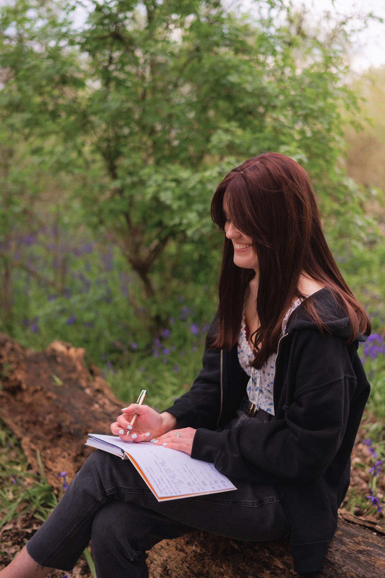 Emily, a young white woman with reddy brown hair, wearing black jeans and a black jumper is sat on a log in the woods holding a pen and looking down at a notebook.