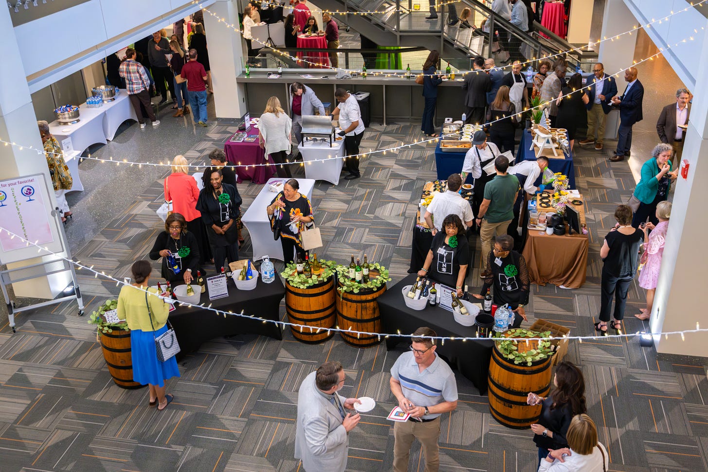 attendees are standing at a variety of wine pouring stations in an open area with lights strung across columns attendees are standing at a variety of wine pouring stations in an open area with lights strung across columns