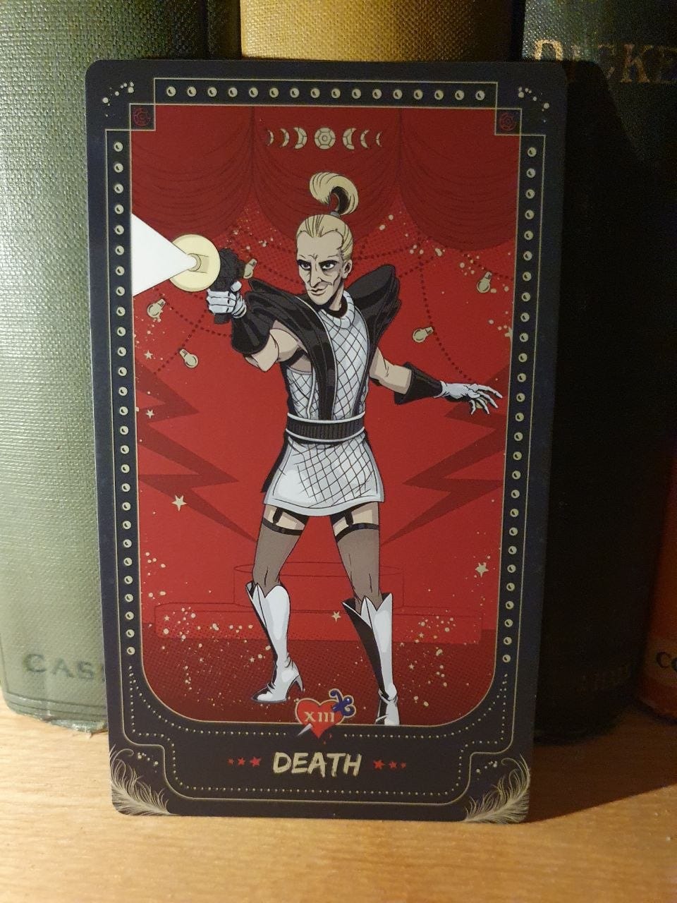 Death tarot card, XIII, depicting a blond figure with a high ponytail, futuristic black-and-white costume and ray gun, set against a dramatic red theatrical background. Death tarot card, XIII, depicting a blond figure with a high ponytail, futuristic black-and-white costume and ray gun, set against a dramatic red theatrical background.