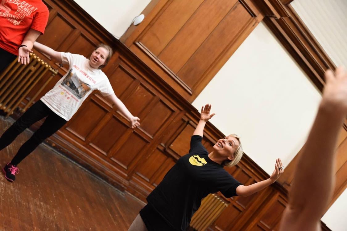 An image of Laura, a middle-aged white woman with short blonde hair, stretching out with some other participants of her company Loop Theatre. They are in a room with wooden panels on the walls.