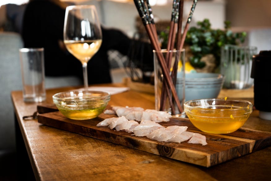 Image shows a wooden plank with pieces of fish it and a glass of wine in the background, with two bowls of fluid to dip the fish in. Image shows a wooden plank with pieces of fish it and a glass of wine in the background, with two bowls of fluid to dip the fish in.