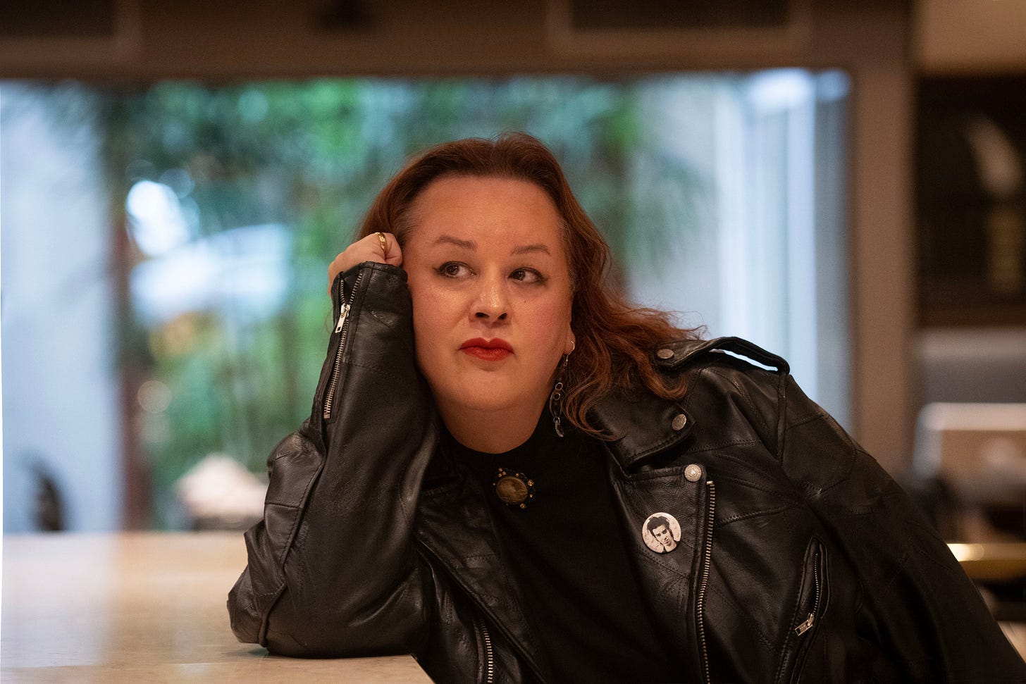 A person with long hair rests their chin on their hand, wearing a black leather jacket and a round pin, in a softly lit indoor setting.