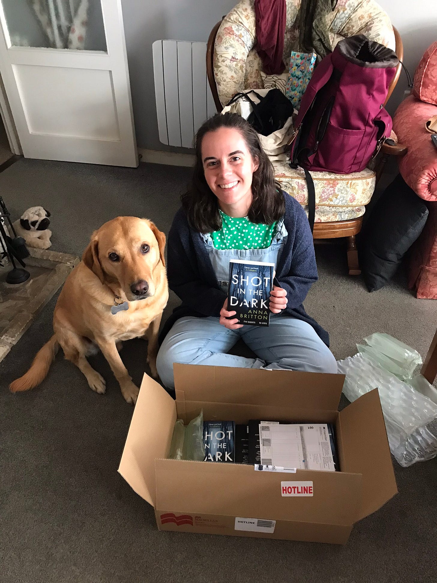 A brown haired white woman sits beside a ginger labrador. They have opened a box containing lots of copies of Shot in the Dark