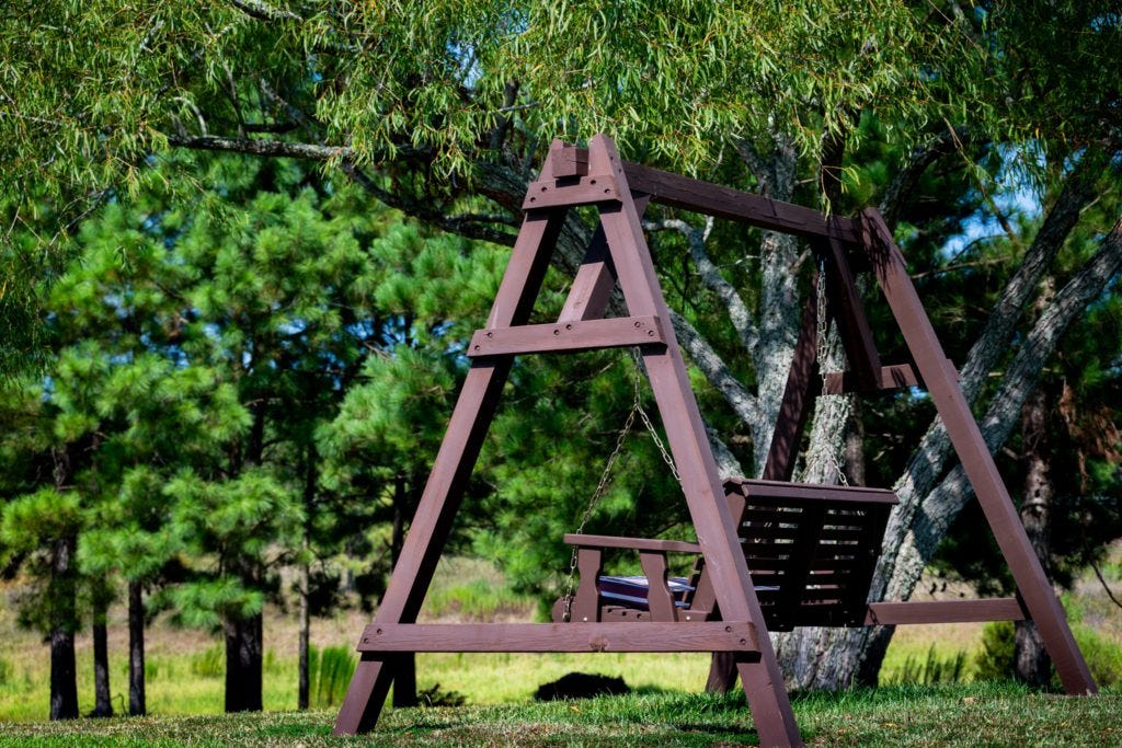 A wooden swing set with a seating swing, shaded by lush trees and surrounded by green grass under a clear blue sky.
