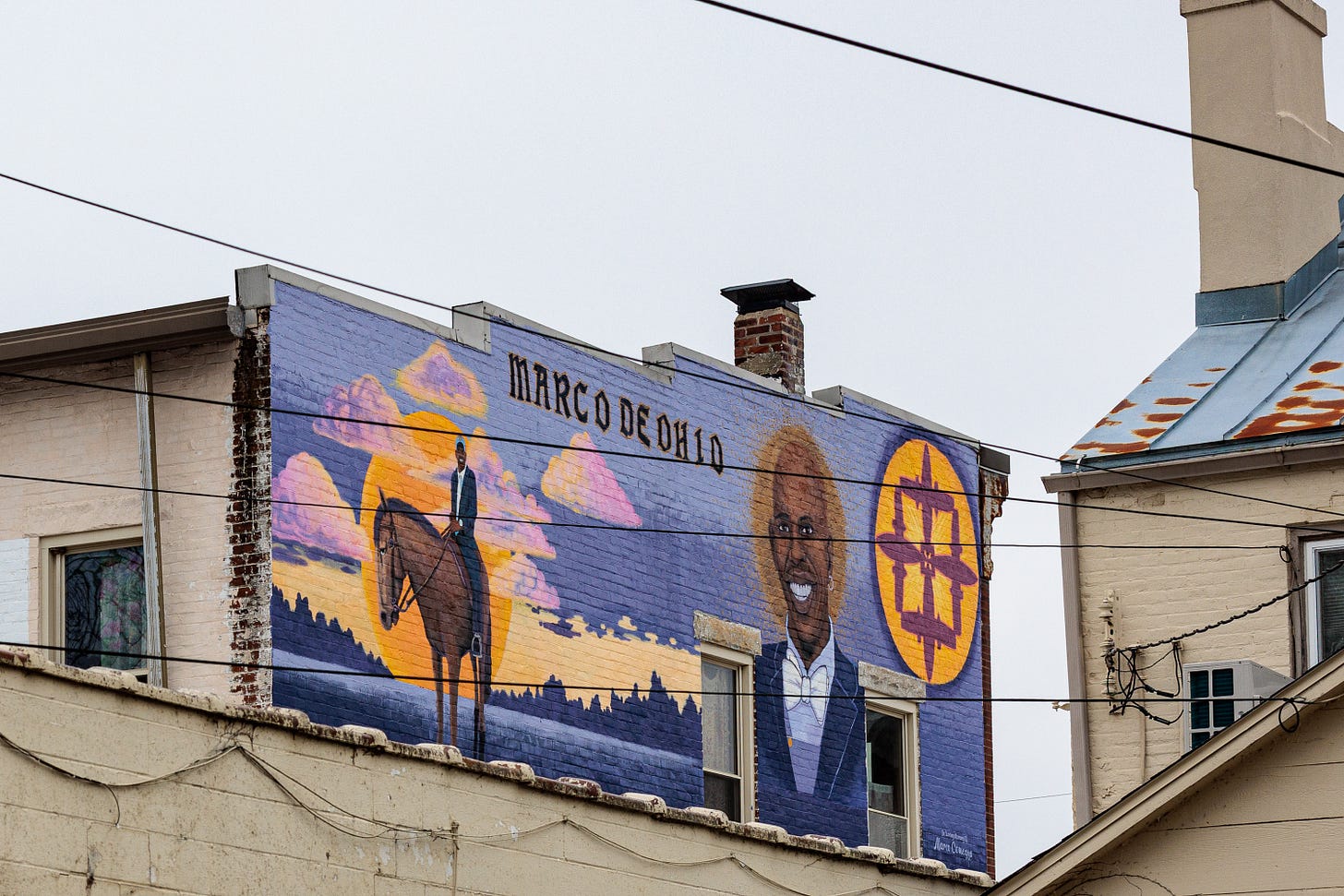 A mural titled 'Marco De Ohio' showing a man riding a horse beside a large portrait of a smiling man, set against a sunset sky.