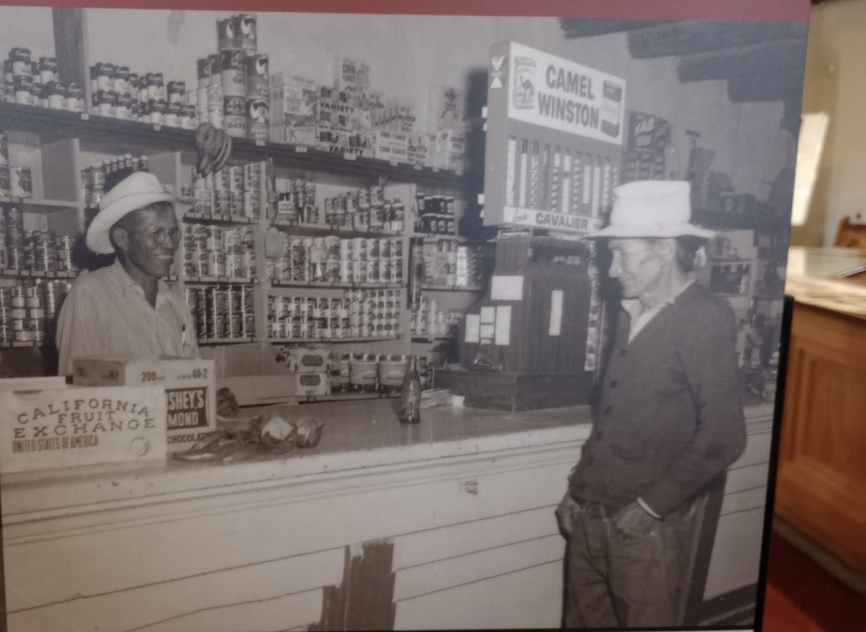 Man in cowboy hat behind a counter, with shelving behind him of canned and packaged groceries, cigarette display, talking to customer in cowboy hat