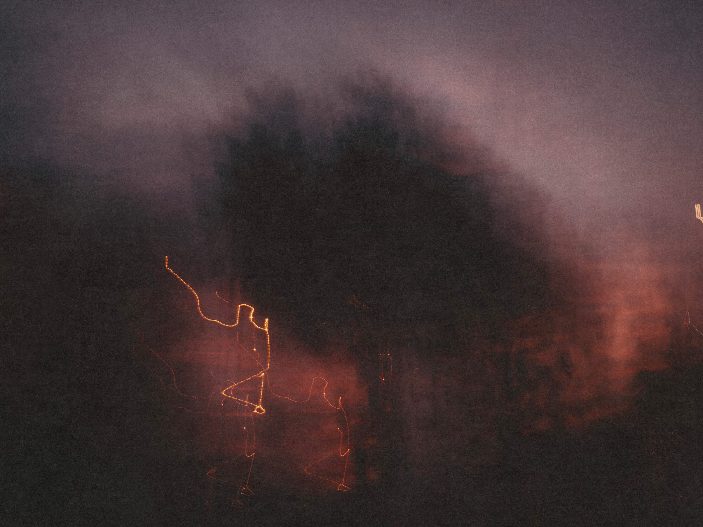 A tree appears in dark silhouette against a dusk sky. The image is soft and blurred, consistent with a hand-held long exposure. In the background, thin light trails form looping, wavy lines near the lower left and centre, with faint warm tones near the horizon.