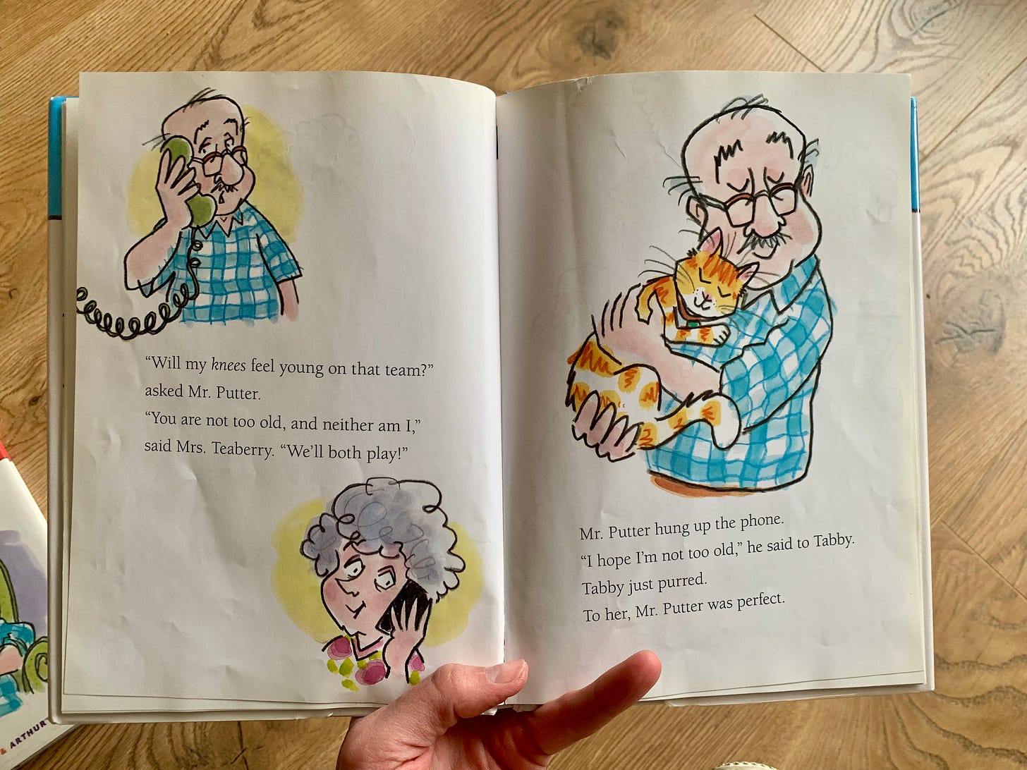 Rebekah holds open one of the Mr Putter and Tabby books. The illustrations on the left page include Mr Putter and Mrs Teaberry talking on their phones. The text captures a conversation between the two. Mr Putter worries he’s too old to play on the sports team. Mrs Teaberry says neither of them are too old. The illustration on the right shows Mr Putter snuggling his cat Tabby. The text reveals Mr Putter is still worried he’s too old, but his cat Tabby snuggles in. She thinks he’s perfect. 
