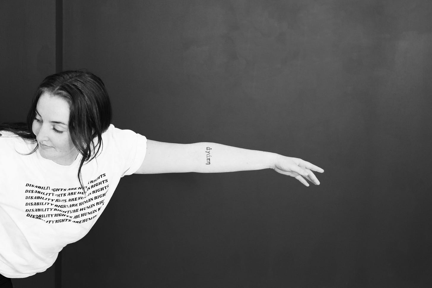 A black and white medium close-up of a woman in a white t-shirt, who looks down to the side, extends her arm with a visible "áɡápē" tattoo, against a dark wall.