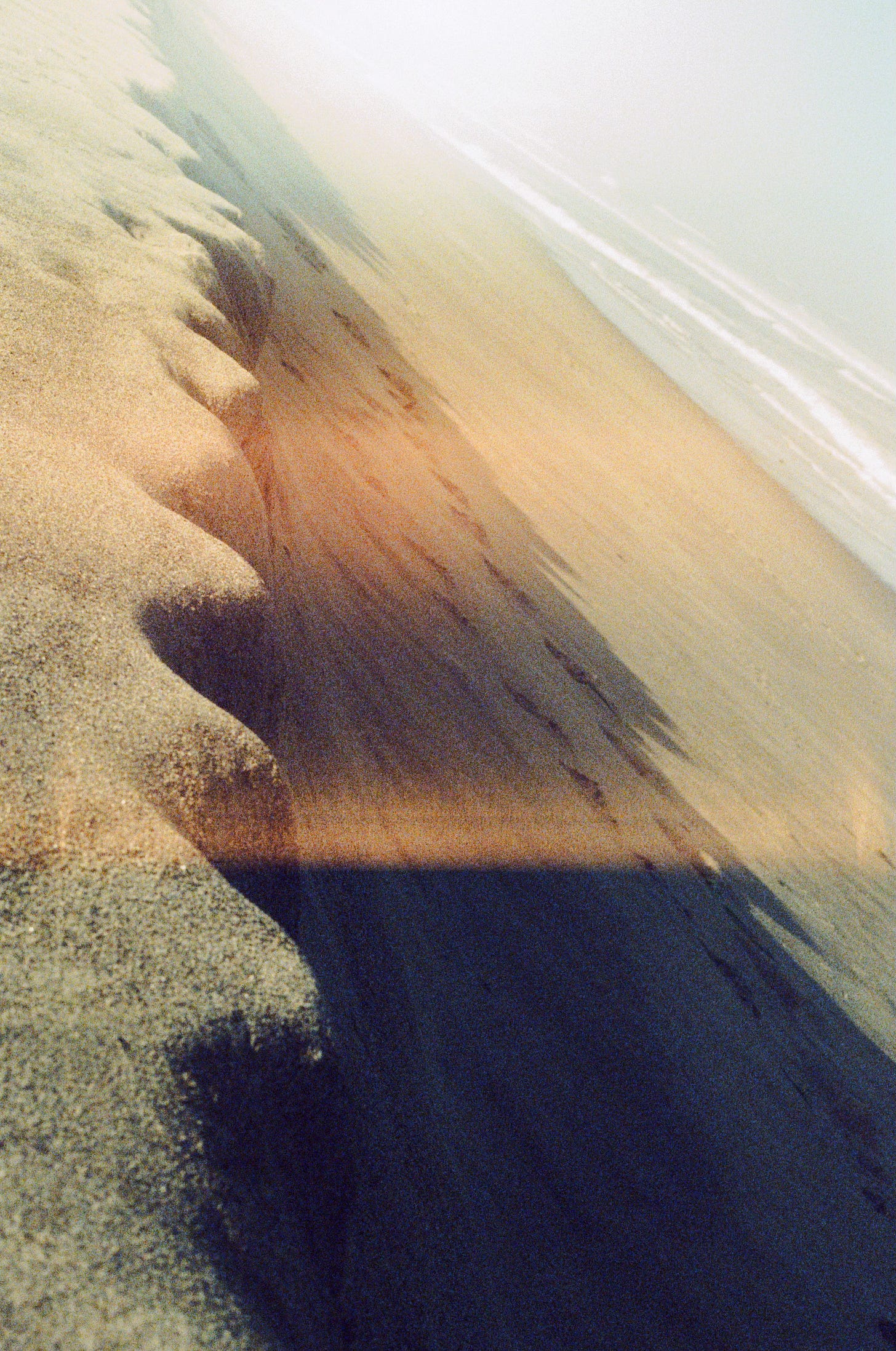 A film photograph of a sand shelf with a wavy edge near the ocean; the water can be seen in the background--in the upper right corner of the frame. Some thin, faint red light leaks cross the lower part of the frame horizontally.