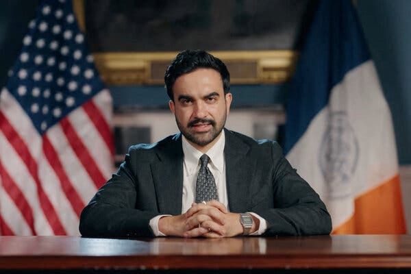 Mayor Zohran Mamdani looks solemnly ahead as he places both forearms and his clenched hands on a desk.