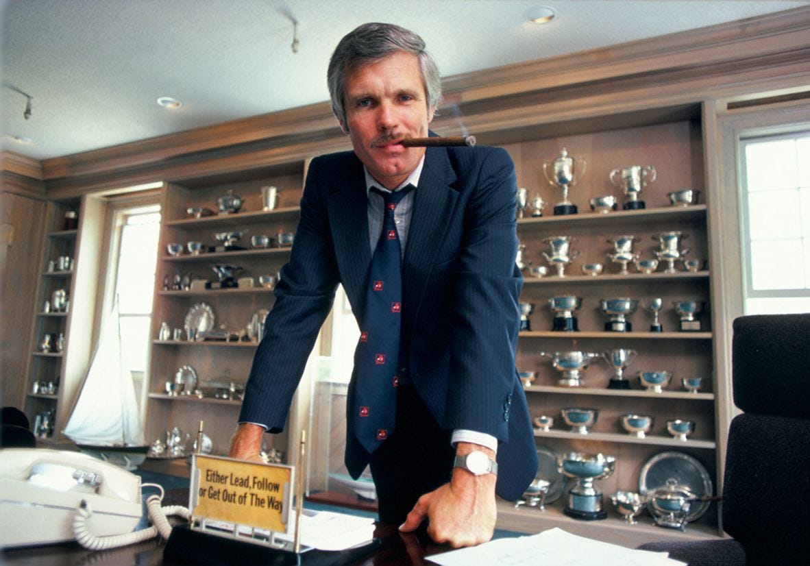 Turner at his desk in 1985. The sign reads "Either Lead, Follow or Get Out of The Way."