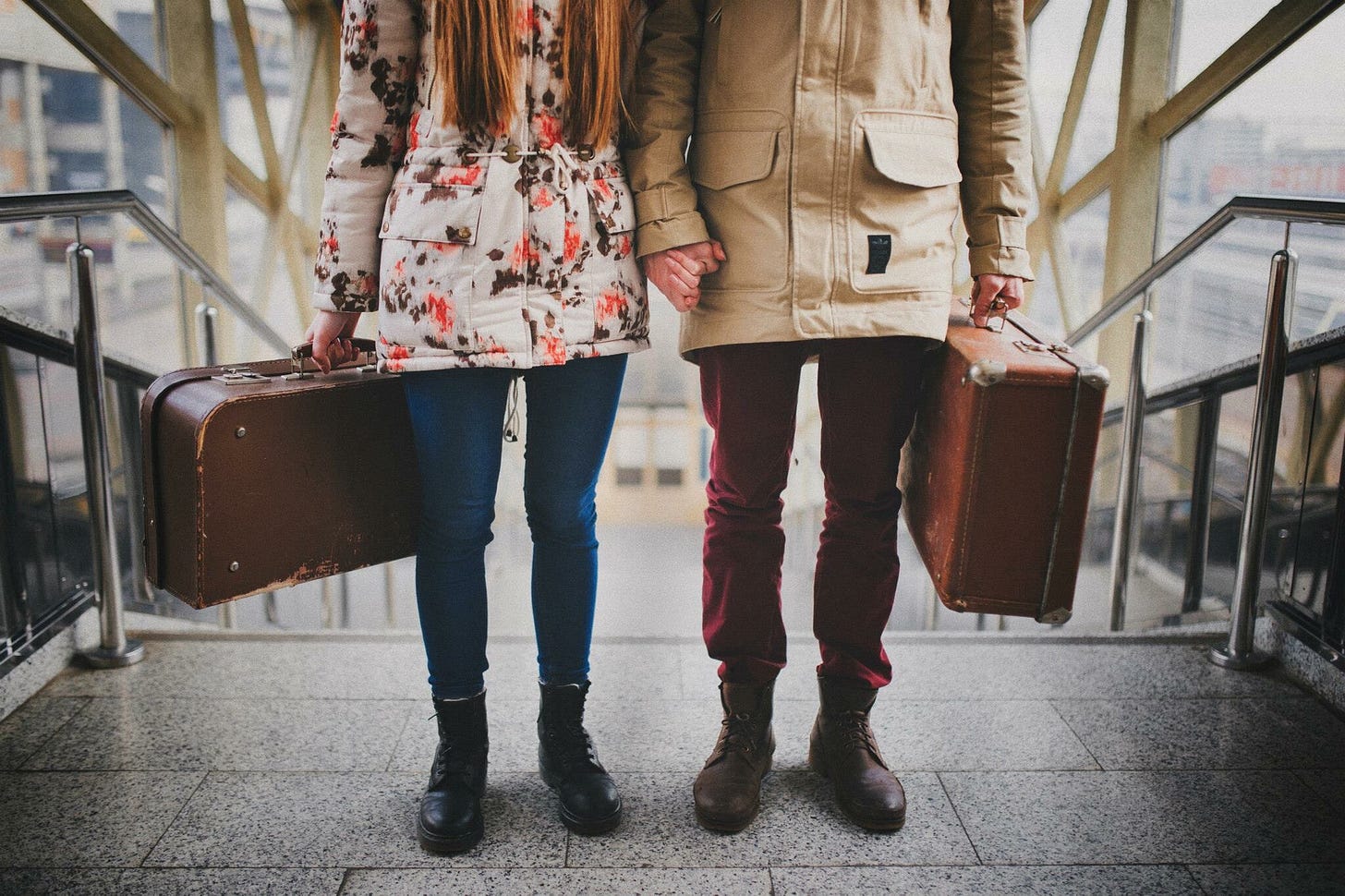 A couple stands holding hands and luggage at the top of the stairs.
