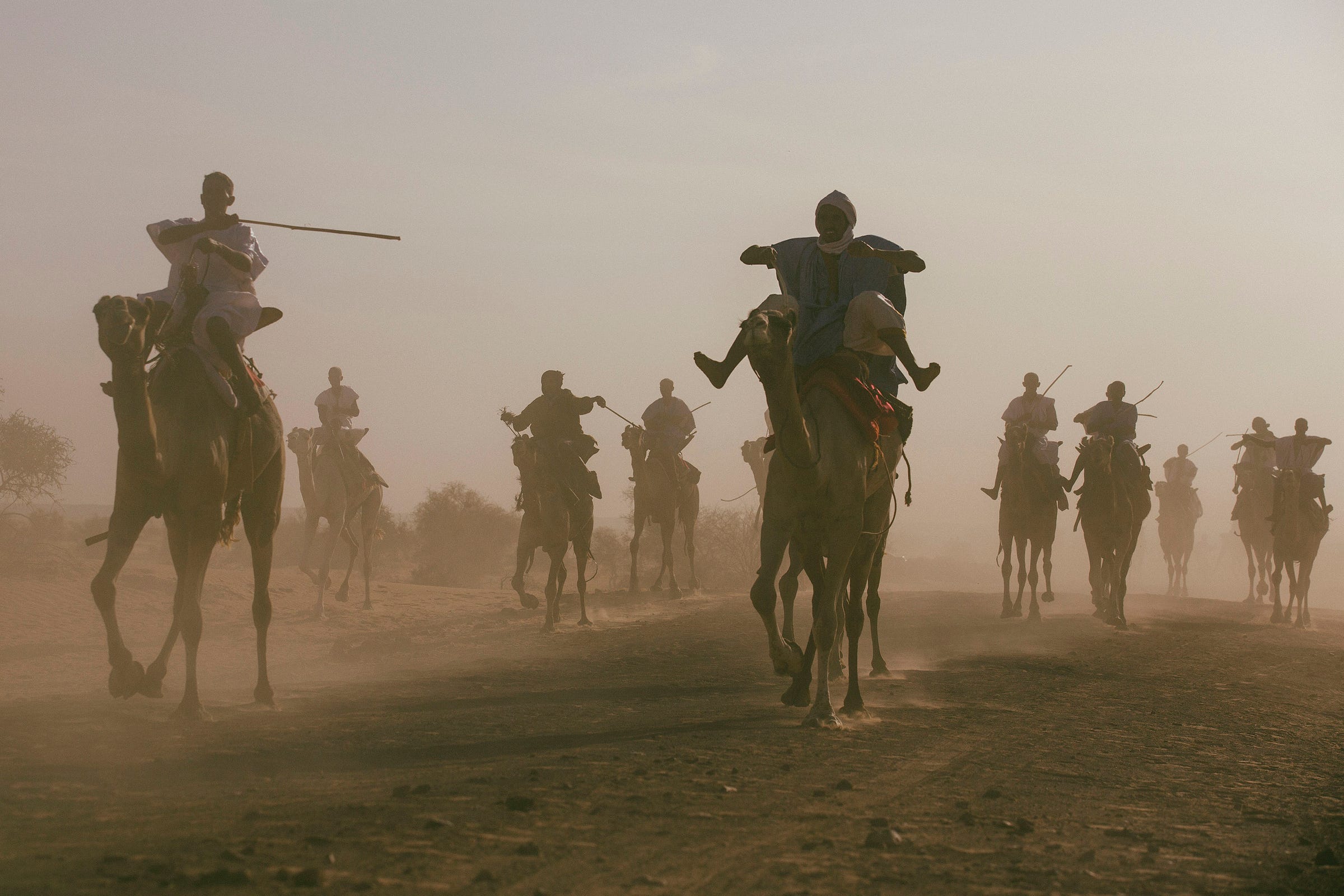 A group of people riding camels through a dusty landscape, some holding spears, with silhouettes visible against the hazy sky. A group of people riding camels through a dusty landscape, some holding spears, with silhouettes visible against the hazy sky.