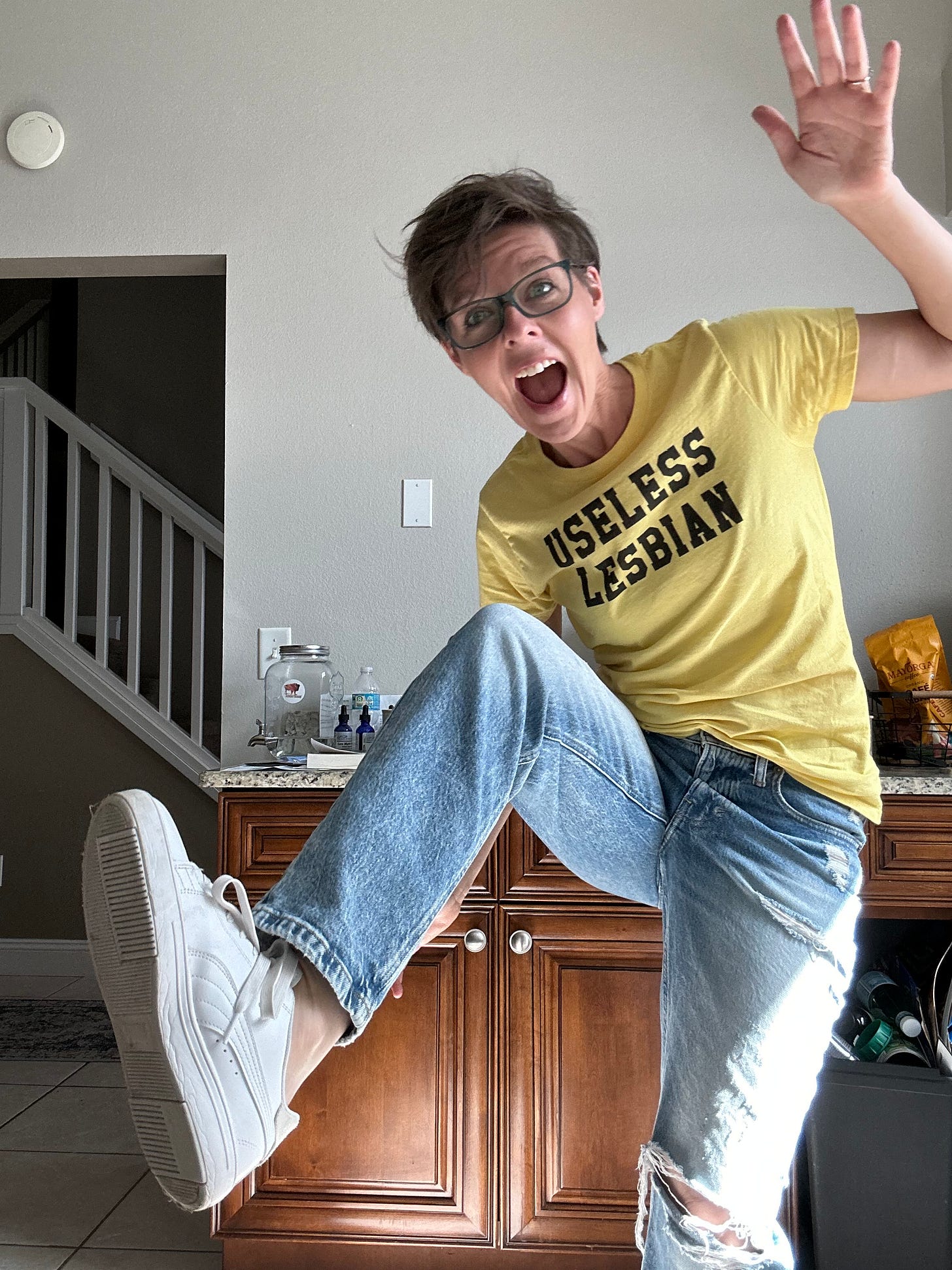 Woman with short brown hair strikes a goofy pose, wearing a bright yellow t-shirt that reads "Useless Lesbian."
