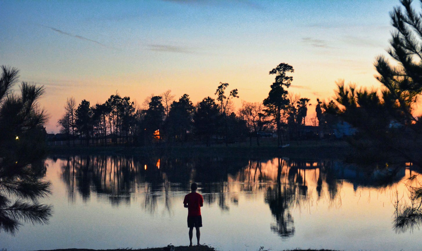 A man facing the sunset over a lake fishing with the reflection of the trees and sky in the lake
