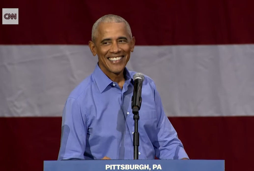 Barack Obama, standing at a lectern in front of a flag-themed backdrop, grins as he gives a speech Barack Obama, standing at a lectern in front of a flag-themed backdrop, grins as he gives a speech