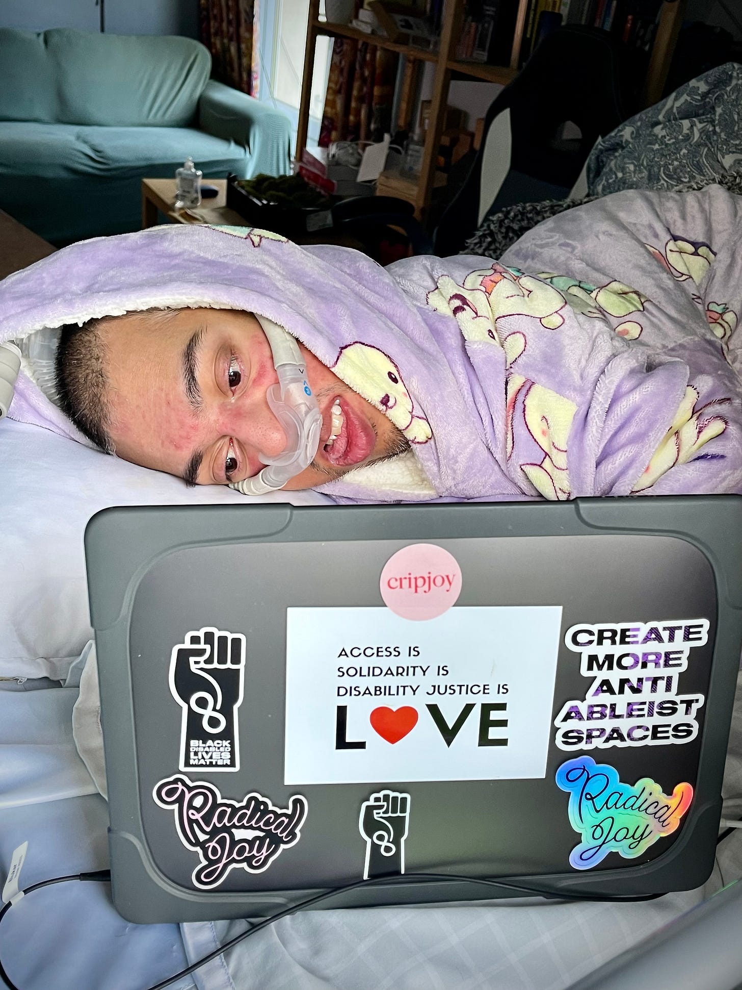 Image description: Bold, wild, fearless existence. In the middle of an abstract dark blue background is a photo of Sulaiman (a wholeheartedly Disabled AF and British-Pakistani man) lying in bed on his right with a nasal ventilator mask over his face as he works on his MacBook Pro laptop. On the grey case of his MacBook Pro are 7 stickers on the back of the screen via Sulaiman himself and his Disabled global kin (by bond) – Jennifer White-Johnson, Alice Wong, Sandy Ho, and Mia Mingus. Sticker 1 is a circular pink sticker with red text that says “Cripjoy.” Sticker 2 is a sticker with a white background featuring a gradient black to purple text that says “Create More Anti-Ableist Spaces” in capital letters. Stickers 3 and 4 are of cursive text that says “Radical Joy,” one a sticker of pink text with a black outline and the other a colourful holographic sticker. Sticker 5 is a graphic of the black power fist against a white background with the infinity symbol and the words “Black Disabled Lives Matter” written in bold white font placed down the wrist of the fist. Sticker 6 is a graphic of the black power fist against a white background with the infinity symbol for neurodiversity in bold white lines placed down the wrist of the fist. Finally, sticker 7 is an extensive rectangular white background with black text over 4 lines; on lines 1 to 3, the text says, “Access is… Solidarity is… Disability Justice is…” and on line 4, the larger text says, “Love” in capital letters with the “o” replaced with a red heart emoji. Sulaiman is wearing his Bunny Oodie (“like a jumper crossed with a blanket crossed with a cloud”) in purple via @the_oodie, and he feels calm as he works on his MacBook Pro. At the top middle of the image is cursive white text that says, “Sulaiman R. Khan.” Below it, in all caps, white text is “Daringly Integrating Disability.” At the bottom right corner of the image is the social logo of ThisAbility Limited in all white featuring the Adinkra symbol for interdependence. End. Image description: Bold, wild, fearless existence. In the middle of an abstract dark blue background is a photo of Sulaiman (a wholeheartedly Disabled AF and British-Pakistani man) lying in bed on his right with a nasal ventilator mask over his face as he works on his MacBook Pro laptop. On the grey case of his MacBook Pro are 7 stickers on the back of the screen via Sulaiman himself and his Disabled global kin (by bond) – Jennifer White-Johnson, Alice Wong, Sandy Ho, and Mia Mingus. Sticker 1 is a circular pink sticker with red text that says “Cripjoy.” Sticker 2 is a sticker with a white background featuring a gradient black to purple text that says “Create More Anti-Ableist Spaces” in capital letters. Stickers 3 and 4 are of cursive text that says “Radical Joy,” one a sticker of pink text with a black outline and the other a colourful holographic sticker. Sticker 5 is a graphic of the black power fist against a white background with the infinity symbol and the words “Black Disabled Lives Matter” written in bold white font placed down the wrist of the fist. Sticker 6 is a graphic of the black power fist against a white background with the infinity symbol for neurodiversity in bold white lines placed down the wrist of the fist. Finally, sticker 7 is an extensive rectangular white background with black text over 4 lines; on lines 1 to 3, the text says, “Access is… Solidarity is… Disability Justice is…” and on line 4, the larger text says, “Love” in capital letters with the “o” replaced with a red heart emoji. Sulaiman is wearing his Bunny Oodie (“like a jumper crossed with a blanket crossed with a cloud”) in purple via @the_oodie, and he feels calm as he works on his MacBook Pro. At the top middle of the image is cursive white text that says, “Sulaiman R. Khan.” Below it, in all caps, white text is “Daringly Integrating Disability.” At the bottom right corner of the image is the social logo of ThisAbility Limited in all white featuring the Adinkra symbol for interdependence. End.